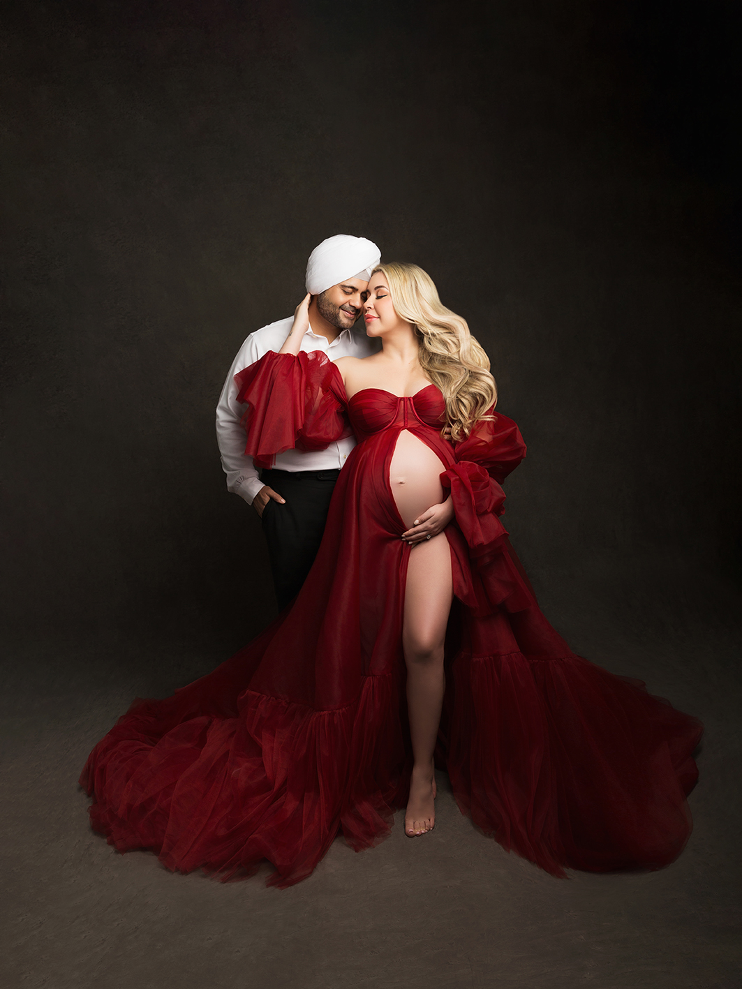 XIAOYUN PHOTOGRAPHY Sikh couple luxury maternity shoot — pregnant woman in red gown with exposed belly, husband in white turban, studio portrait.