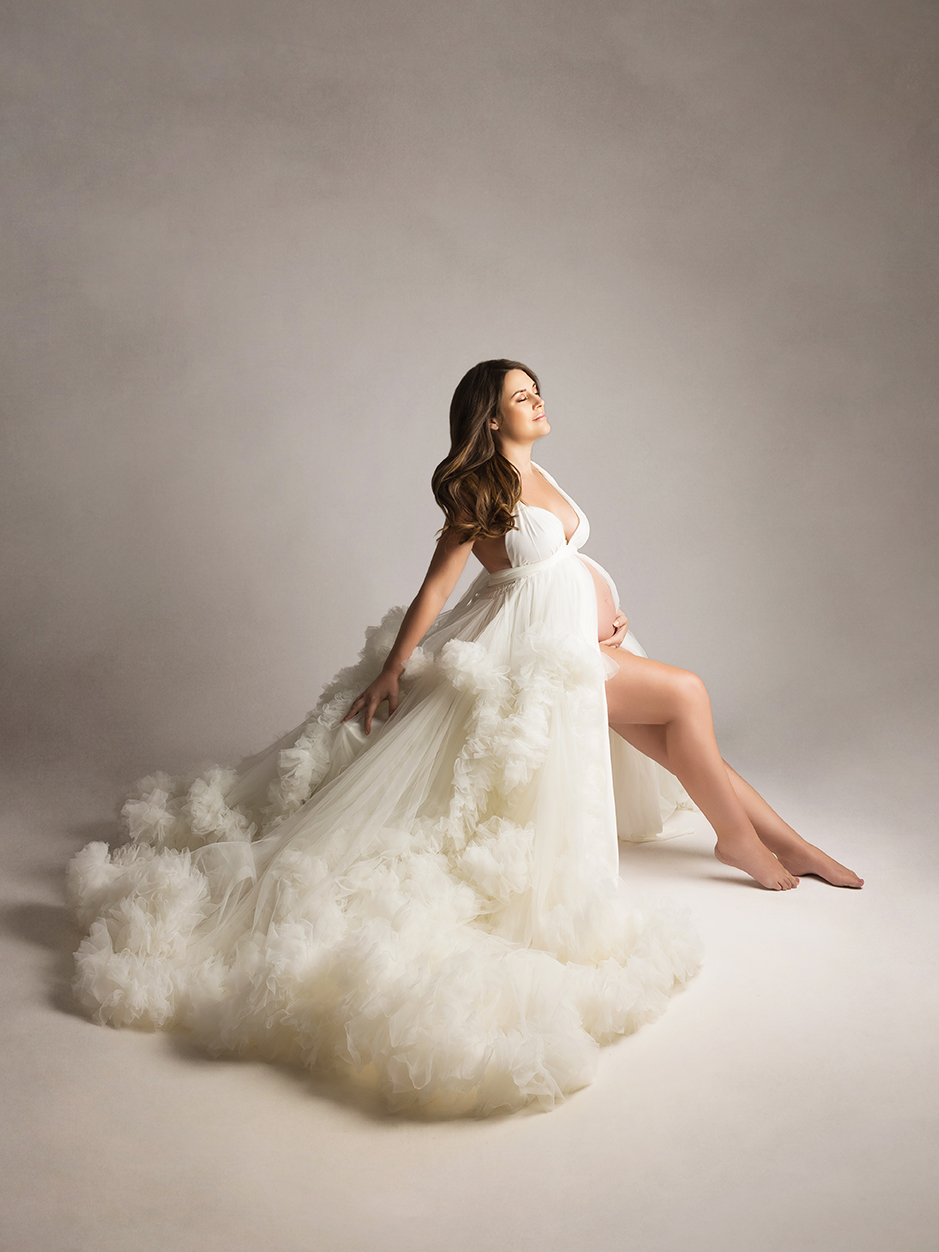 XIAOYUN PHOTOGRAPHY Pregnant woman in white tulle gown with ruffles, seated in soft glam studio maternity shoot with timeless style