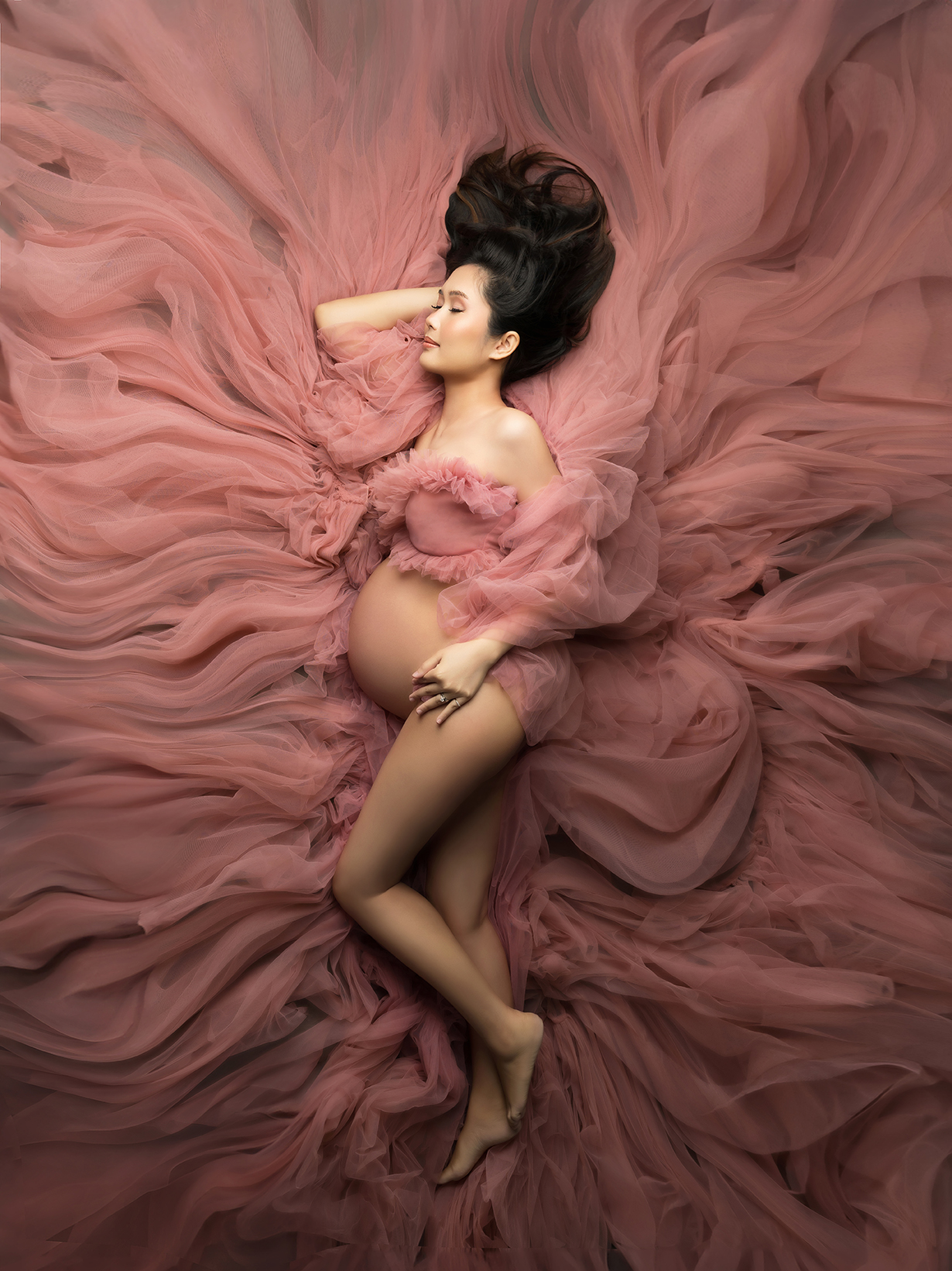 XIAOYUN PHOTOGRAPHY Dreamy soft glam maternity shoot — pregnant woman lying on flowy blush tulle fabric in fine art studio setup